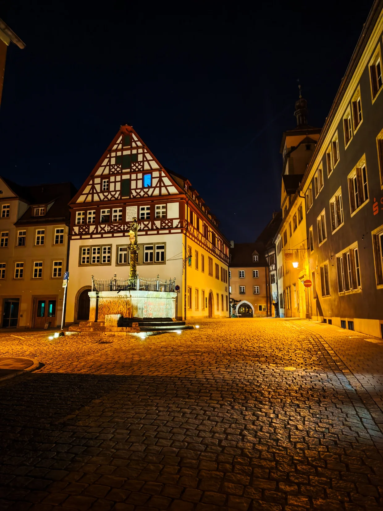 An empty cobbled Rothenburg square at night, a column fountain lit from below