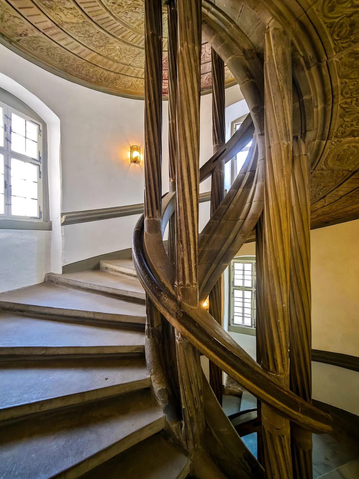A spiral staircase with bundled-stone handrails twisting up under a gilded painted ceiling