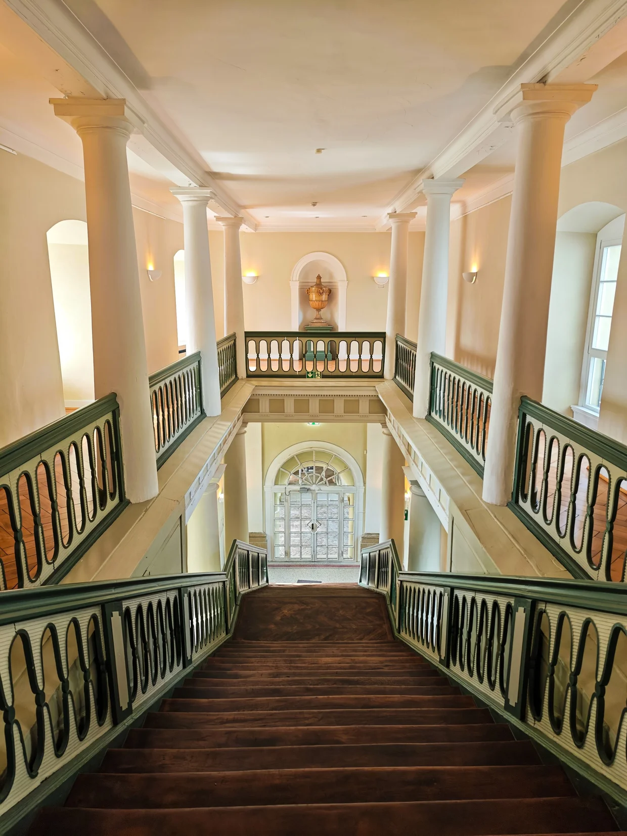 A wide neoclassical staircase with plain columns, green-and-white balustrades and an urn in a niche at the top