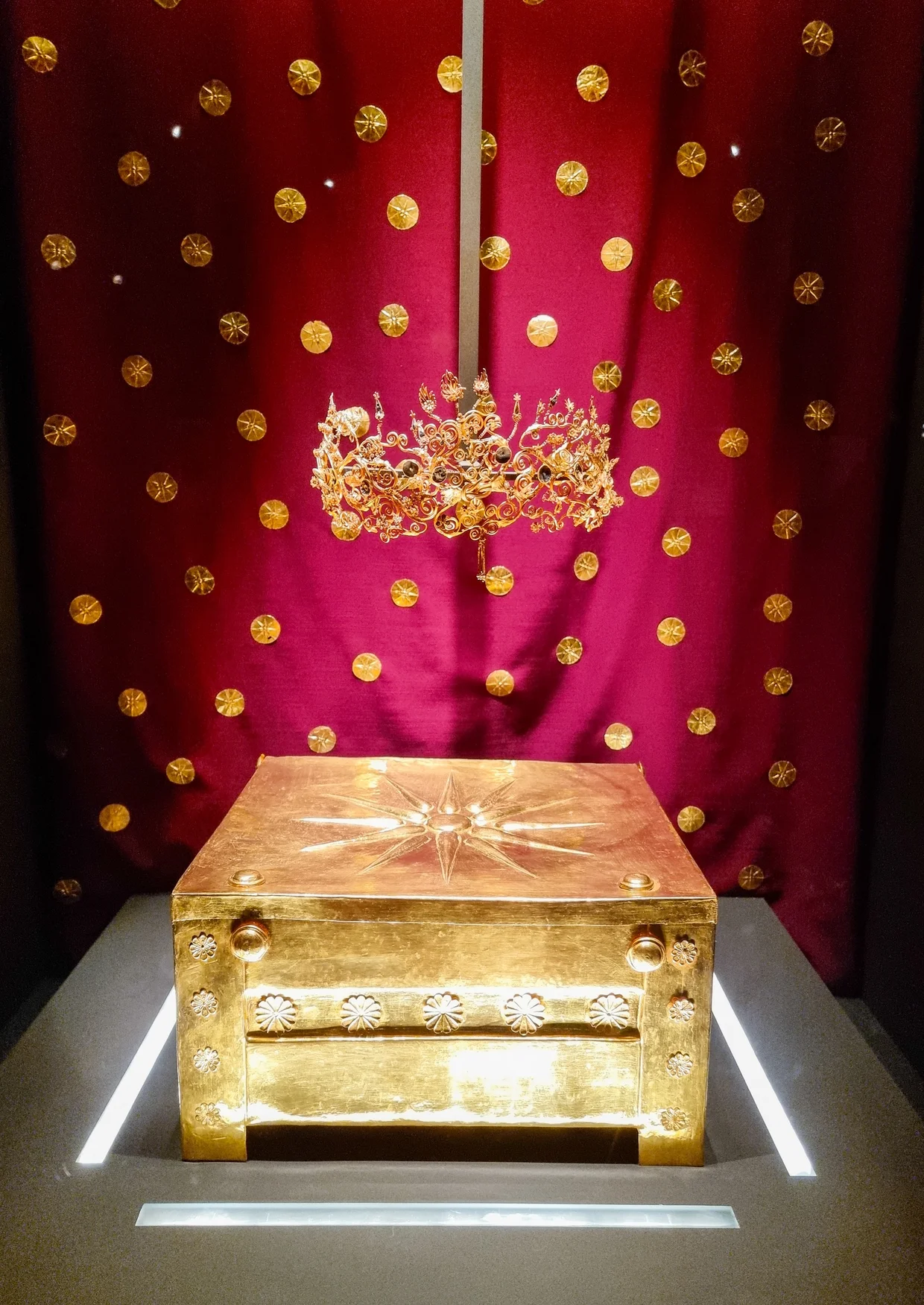 Gold larnax embossed with the Vergina star, set against a red velvet backdrop hung with gold rosettes
