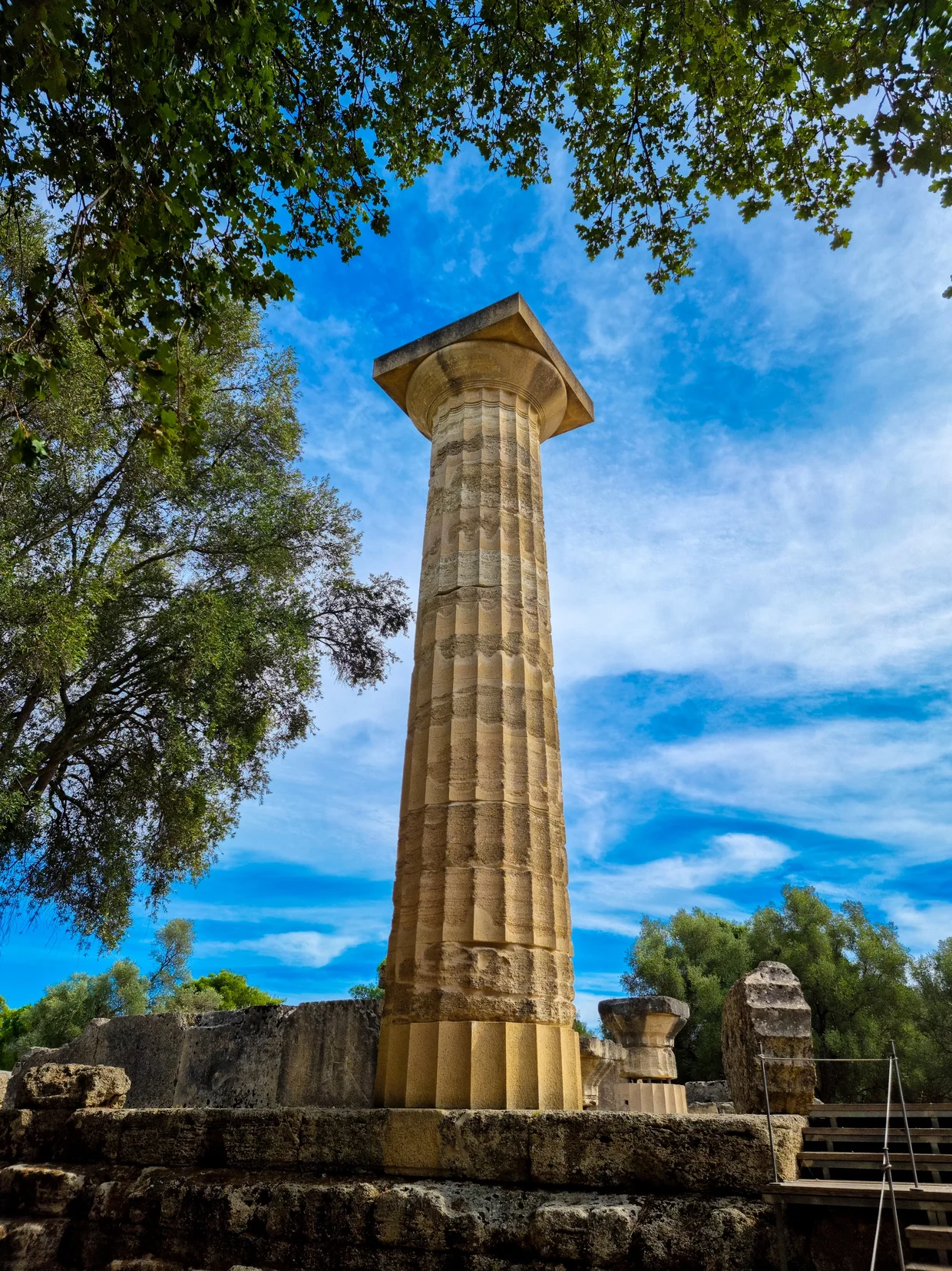 A single re-erected Doric column from the Temple of Zeus at Olympia, framed by trees against a blue sky