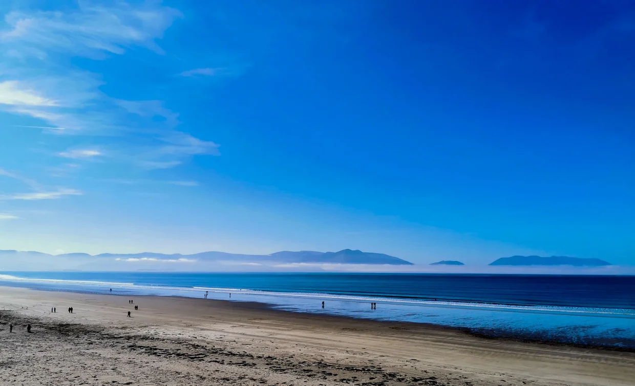 A wide low-tide beach on the Kerry coast with distant walkers and hazy mountains