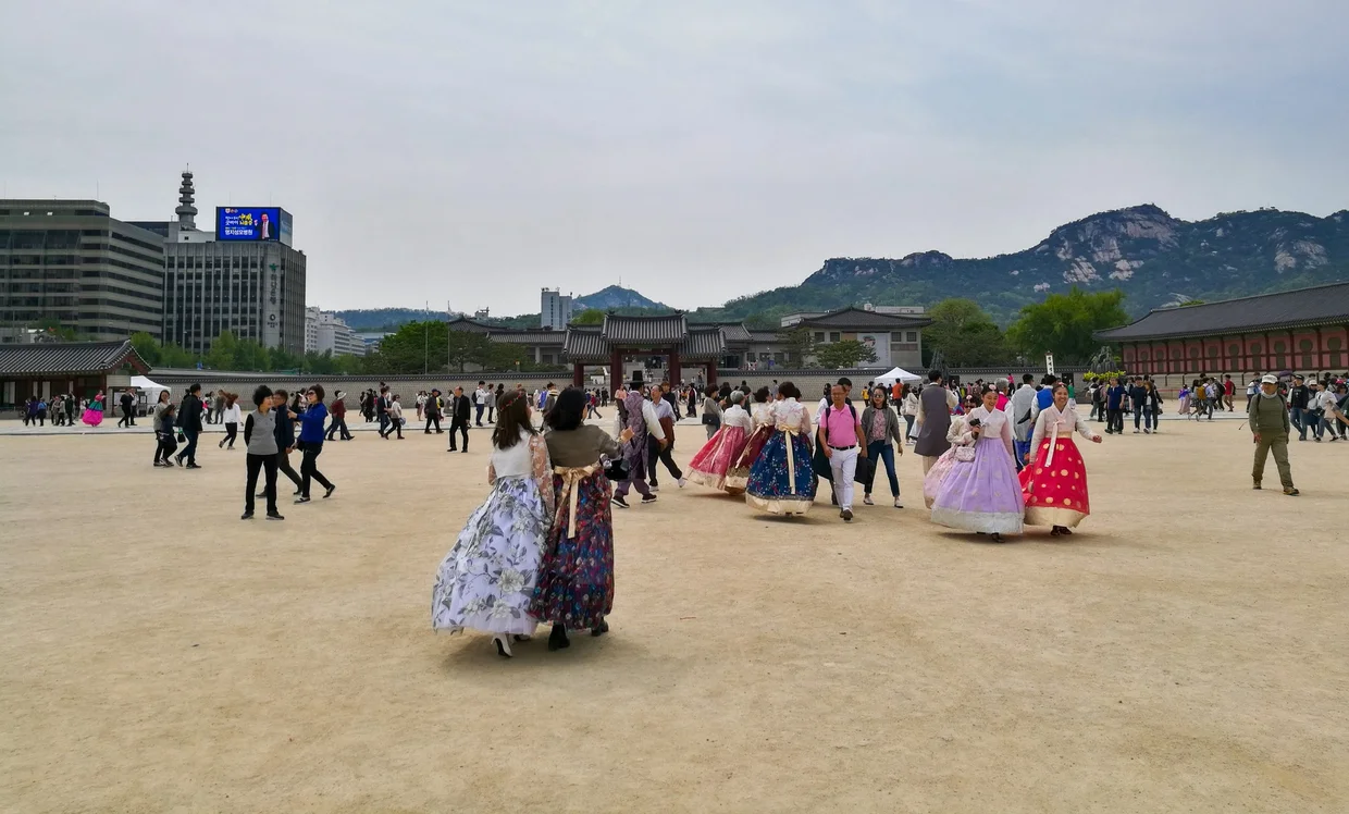 Wide palace grounds with visitors in colorful hanbok, mountains rising behind